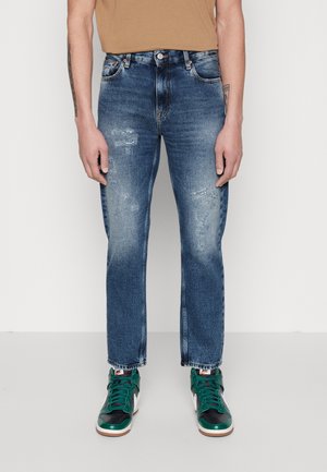 Homme portant un jean slim bleu délavé avec une légère usure et des baskets montantes vertes et noires, debout sur un fond blanc.