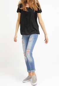 Czarny krótki top, jasnoniebieskie dżinsy z przetarciami i szare sneakersy wsuwane; casualowy strój na jednolitym tle.