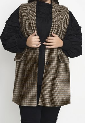 Person med sort bluse og puffede ærmer iført brun og hvid hundetandsvest, der holder vestens revers med begge hænder.