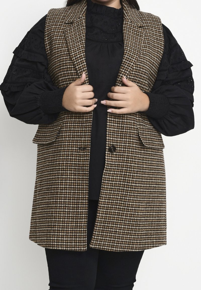 Personne portant un chemisier noir à manches bouffantes et un gilet à motif pied-de-poule marron et blanc, tenant les revers du gilet avec les deux mains.