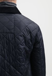 Marineblå kviltad jacka med en diamantmönster, slät textur, manchesterkrage och diskreta sömnadsdetaljer på baksidan.