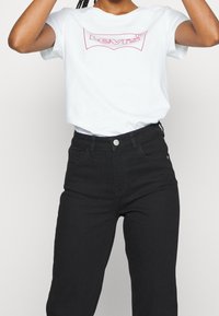 Bílý tričko s kulatým výstřihem a červeným logem Levi's, spojené s vysokým černým džínem s předním zapínáním na knoflík a bočními kapsami.