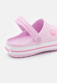 Różowe klapki Crocs wykonane z lekkiego materiału, z białą i różową paskowaną podeszwą. Posiadają tylną pasek oraz mały detal z logo.