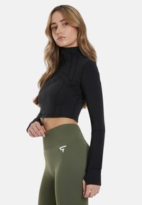 Mustat lyhyet vetoketjulliset takit, joissa on korkea kaulus ja pitkät hihat, yhdistettynä korkeavyötäröisiin oliivinvihreisiin legginsseihin, joissa on valkoinen logo.