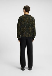 Uomo che indossa pantaloni neri e un maglione a maniche lunghe con un motivo a righe marrone e verde scuro, decorato con motivi di uccelli e fiori, in piedi di spalle su uno sfondo neutro.