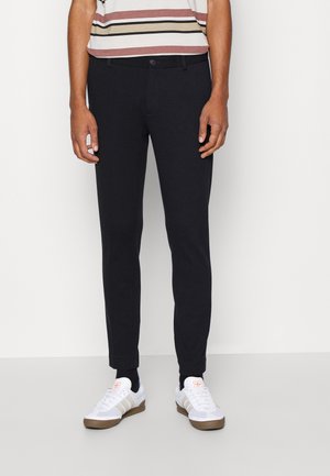 Homme portant un pantalon noir slim, des baskets blanches avec des rayures beiges et une chemise rayée à manches courtes, debout sur un fond blanc.
