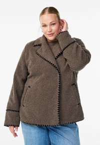 Woman wearing a brown textured fleece jacket with black trim, light blue jeans, and touching her ear while facing forward.