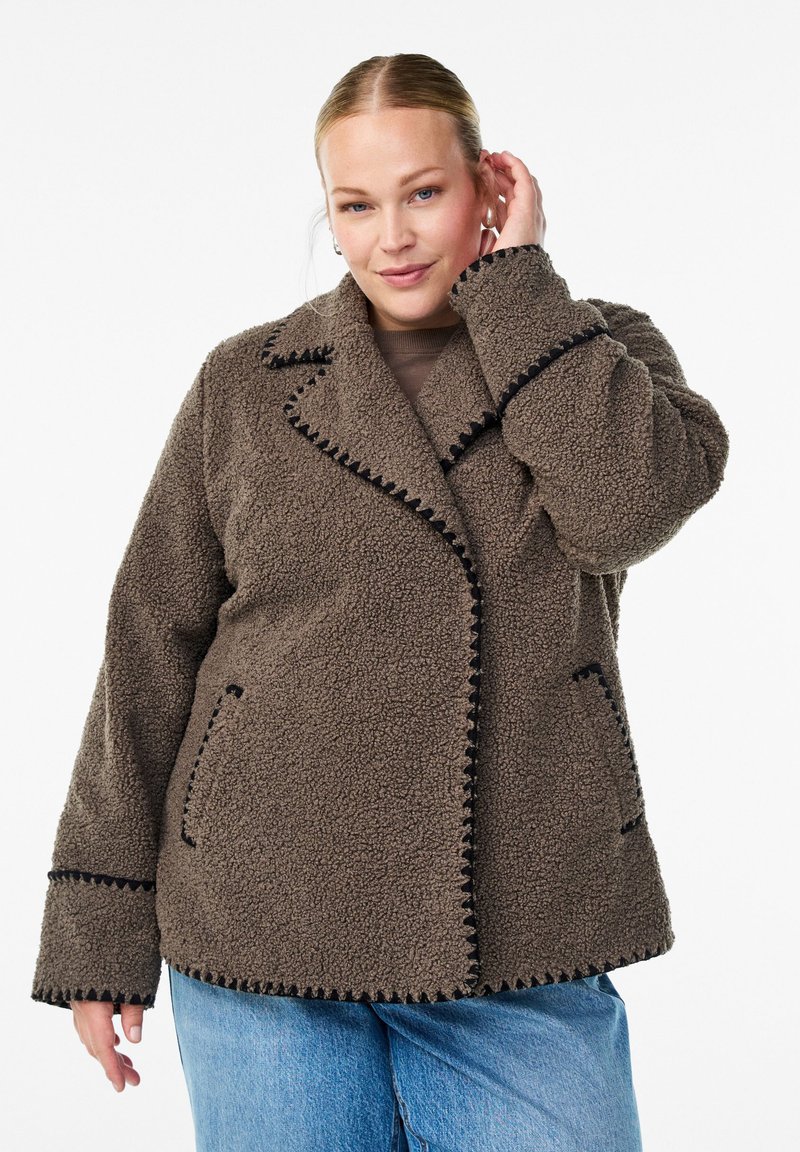 Woman wearing a brown textured fleece jacket with black trim, light blue jeans, and touching her ear while facing forward.