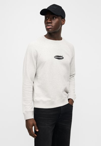 Light gray sweatshirt with raglan sleeves, featuring a black logo patch in the center. Paired with dark jeans and a black cap.