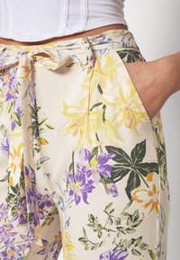 Osoba oblečená v pasu vysokých květinových kalhotách s žlutými a fialovými květinami a zelenými listy, svázaných látkovým pásem, s jednou rukou v kapse.