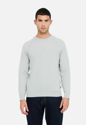 Man wearing a light gray long-sleeve sweatshirt and dark blue jeans, standing facing forward against a plain white background.