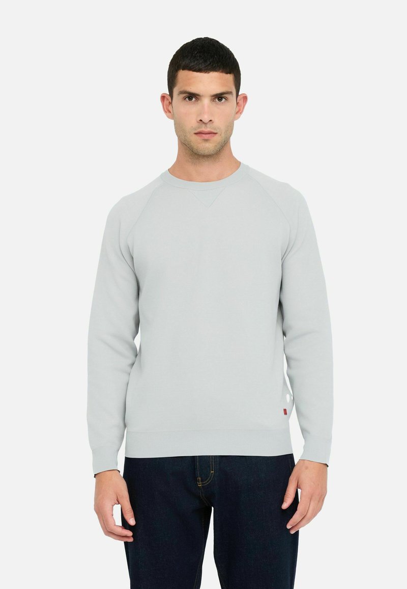 Ein Mann trägt einen hellgrauen Langarm-Pullover und dunkblaue Jeans, steht frontal vor einem schlichten weißen Hintergrund.