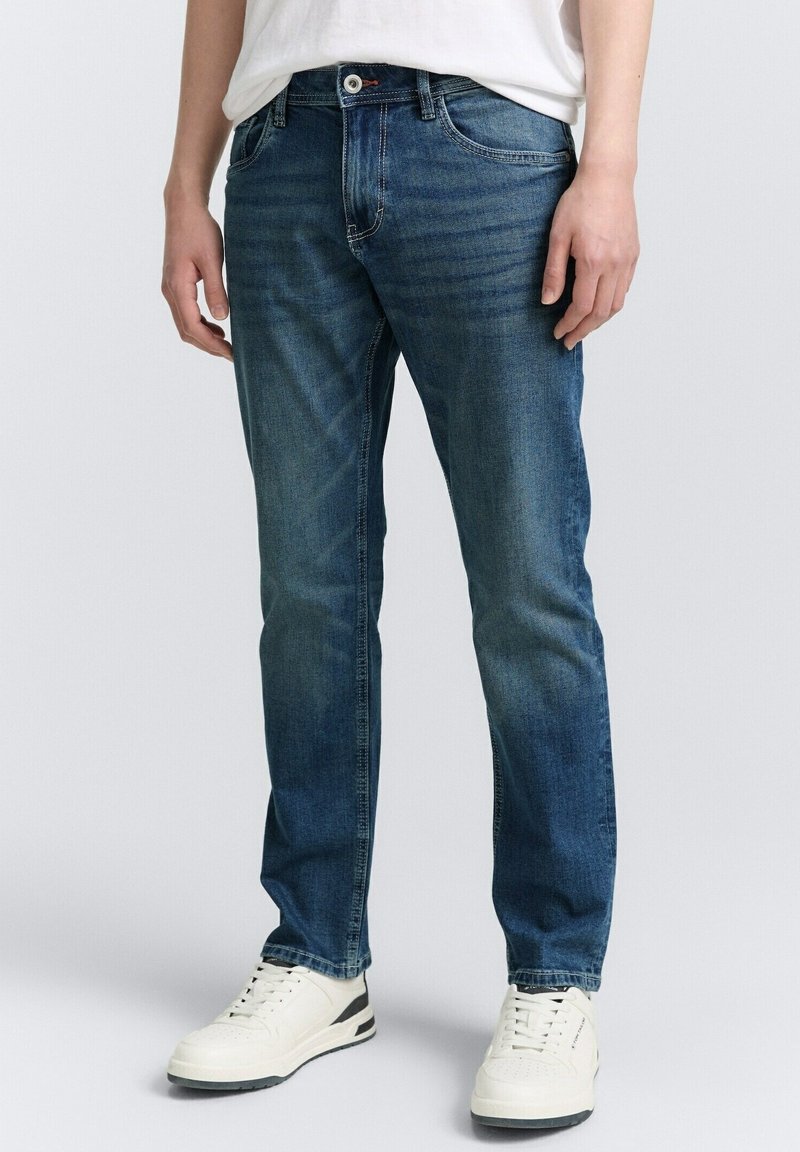 Homme portant un jean bleu slim et des baskets blanches, debout devant un fond uni clair.