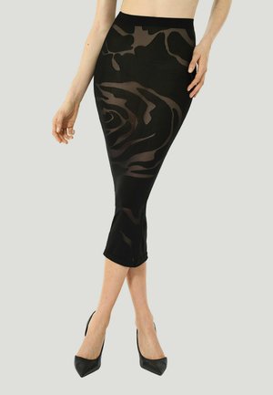 Frau trägt einen schwarzen, hoch taillierten, eng anliegenden Rock mit transparenten Blumenmustern und schwarzen spitzen Schuhen und steht vor einem schlichten Hintergrund.
