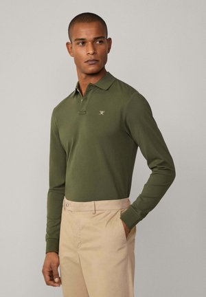 Olijfgroene polo met lange mouwen en een kraag, voorzien van een klein geborduurd logo op de borst. Draagt in combinatie met beige op maat gemaakte broeken.
