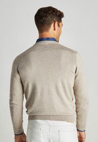 Beige Strickpullover mit rundem Ausschnitt, gerippten Bündchen und Saum. Darunter befindet sich ein blauer Hemdkragen. Kombiniert mit weißen Hosen.