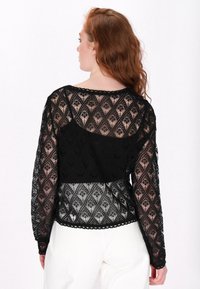 Bluză din dantelă neagră cu mâneci lungi, având un model în formă de diamant, decolteu cu margini zimțate și material semi-transparent. Modelul este prezentat din spate.