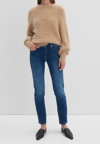 Personne portant un pull beige duveteux, un jean bleu slim-fit et des mocassins noirs pointus, debout devant un fond clair uni.