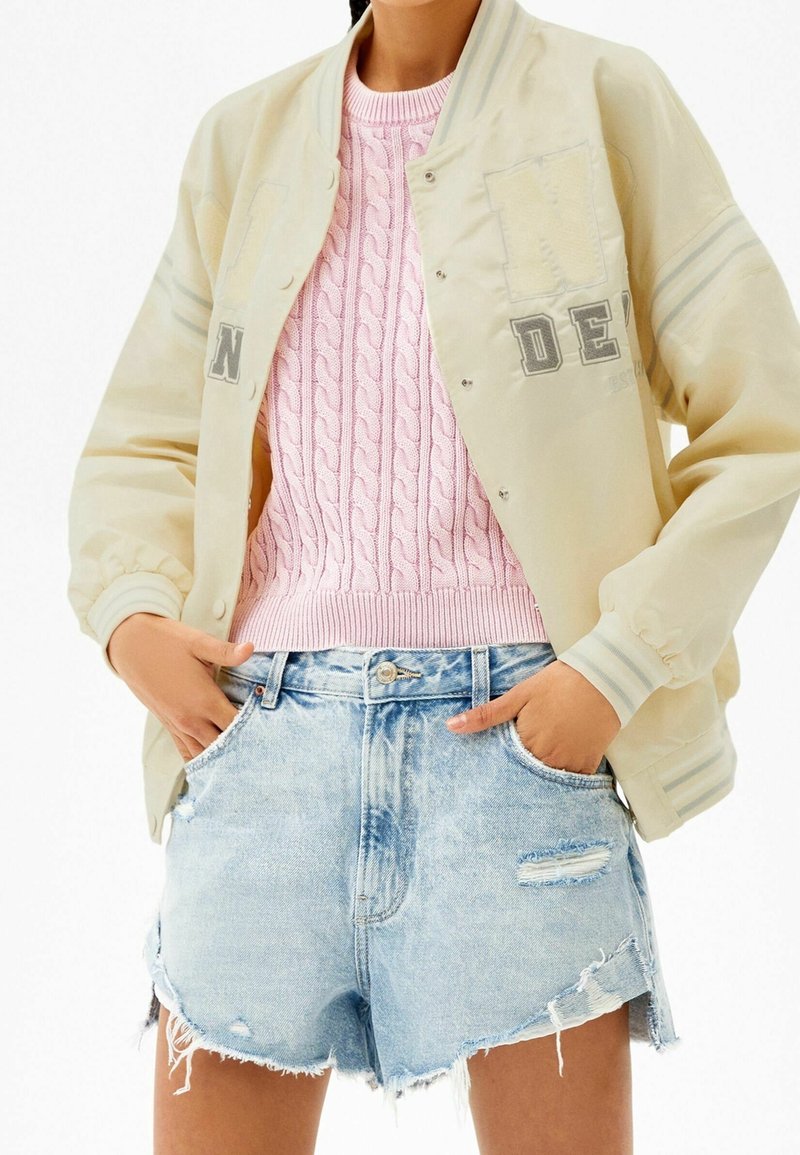 Personne portant une veste universitaire crème par-dessus un pull rose en maille torsadée et un short en jean bleu clair déchiré, les mains dans les poches.