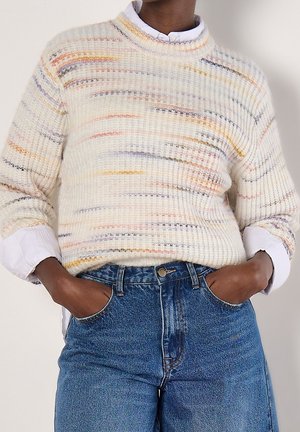 Person trägt einen weißen, strukturierten Pullover mit mehrfarbigen horizontalen Streifen, weiße Hemdärmeln sichtbar und blaue, hoch taillierte Jeans mit den Händen in den Taschen.
