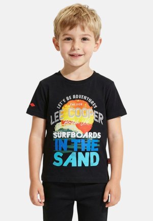 Jeune garçon blond souriant, portant un t-shirt noir avec un texte coloré "Lee Cooper Surfboards In The Sand" et un motif de soleil et de planche de surf.