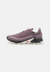 Zapatilla deportiva púrpura con parte superior texturizada, suela de goma, diseño de cordones y acentos en negro y blanco contrastantes. Presenta una puntera reforzada.