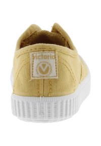 Sneaker di tela gialla con suola in gomma bianca, vista da dietro, che mostra l'etichetta del marchio Victoria sul tallone.