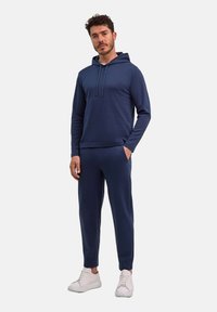 Uomo in piedi con una mano in tasca, indossa un hoodie blu navy e pantaloni abbinati, con scarpe da ginnastica bianche su sfondo bianco.