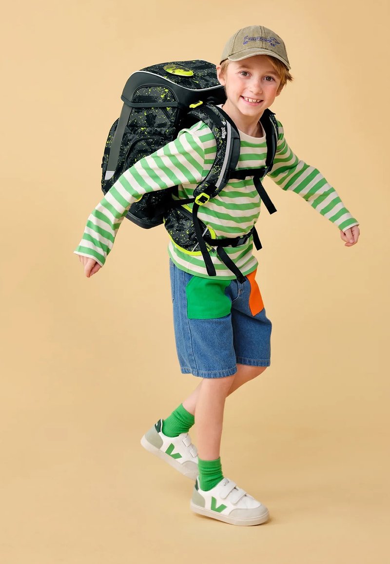 Sac à dos noir avec des accents vert fluo, chemise à rayures vertes et blanches à manches longues, shorts en denim avec des patches colorés, chaussures blanches, chaussettes vertes.