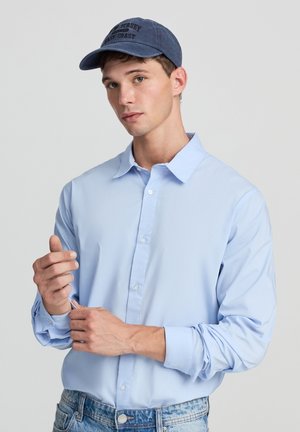 Camicia azzurra chiara a maniche lunghe con colletto appuntito, indossata con un cappellino da baseball blu navy con la scritta ricamata "New Jersey East Coast".
