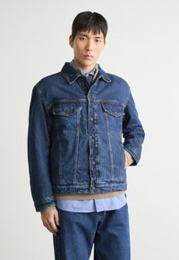 Mörkblå denimjacka med klassisk skärning, som har två bröstfickor, knappstängning och beige ribbad muddar. Bärs över en ljusblå skjorta.