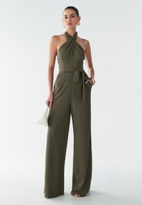 Combinaison vert olive avec un décolleté halter, jambes larges et tissu texturé. Une ceinture assortie se noue à la taille, complétée par un sac à main blanc.