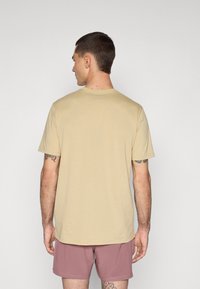 Camiseta de algodón de manga corta color beige, con cuello redondo y corte relajado, mostrada desde atrás, combinada con pantalones cortos marrones y tatuajes visibles.