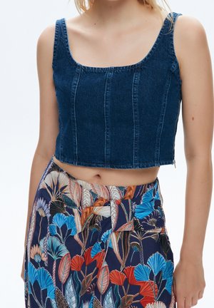 Kobieta w bezrękawnikowym ciemnoniebieskim dżinsowym crop topie i wysokim stanem w spodniach z kolorowym kwiatowo-liściastym wzorem.