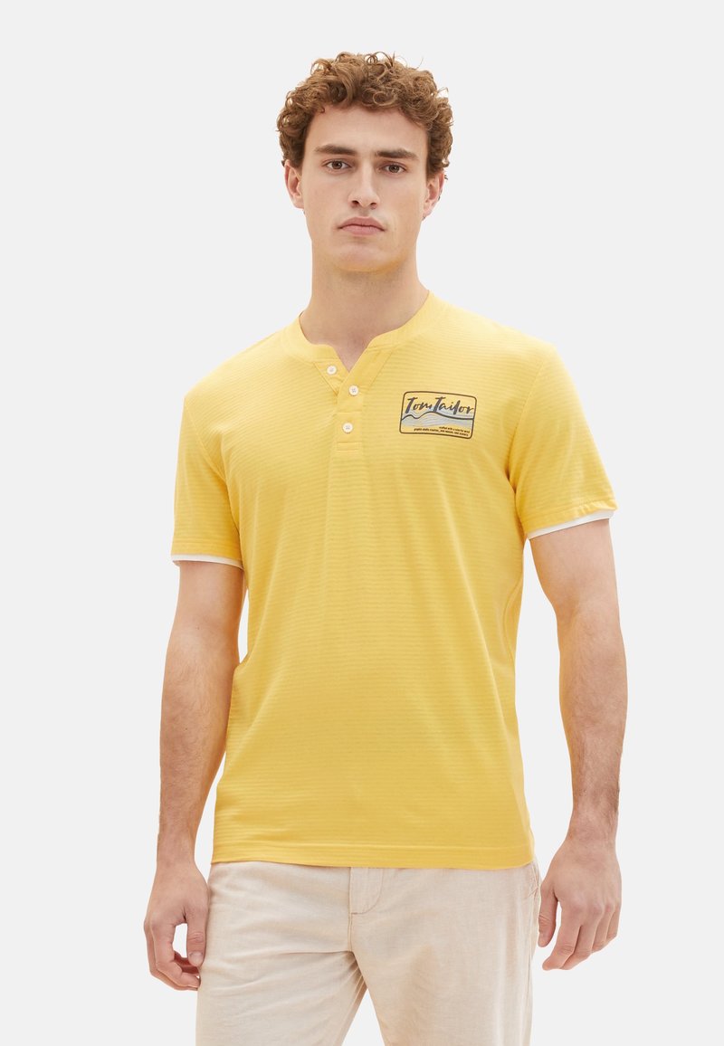 Camisa Henley amarilla de manga corta con cuello tipo polo, tres botones y un pequeño parche de logo. Presenta acentos blancos en las mangas.