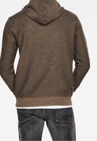 Olivově zelená mikina s kapucí z texturované tkaniny, s žebrovaným lemem a manžetami, s malou značkovou cedulkou poblíž spodní části zad.