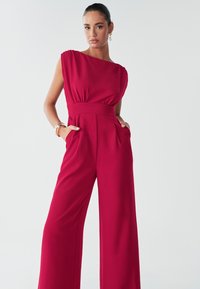Červený overal s jednokrkním střihem, širokými nohavicemi a přiléhavým pasem, vyrobený z lehké látky, s kapsami a záhyby.