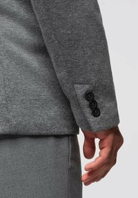 Grå blazer med ett texturerat tyg, med detalj av tre-knappars manschetter och en diskret ficka på ärmen.