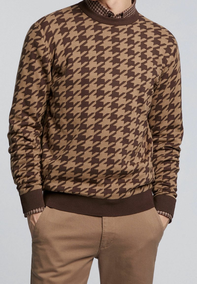 Pull marron et beige à motif pied-de-poule porté sur une chemise à motifs, associé à un pantalon beige, mains décontractées dans les poches, fond neutre.