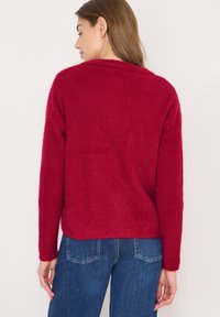 Roter, gerippter Pullover mit rundem Ausschnitt und langen Ärmeln. Weiche Textur, lässige Passform und die Länge bedeckt die Taille, kombiniert mit blauen Jeans.