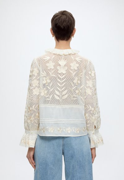 Personne aux cheveux courts portant un chemisier blanc transparent avec broderie florale et poignets à volants, assorti à un jean bleu clair, vue de dos.