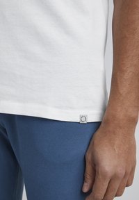 Camiseta de algodón blanca con un dobladillo redondeado, que presenta una pequeña etiqueta en el lado inferior. Combinada con pantalones azules texturizados.