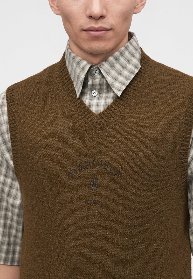Mann trägt eine braune gestrickte V-Ausschnitt-Weste über einem grauen karierten Hemd mit Knöpfen und Kragen, blickt nach vorne, zugeschnitten am Kinn und in der Mitte des Oberkörpers.