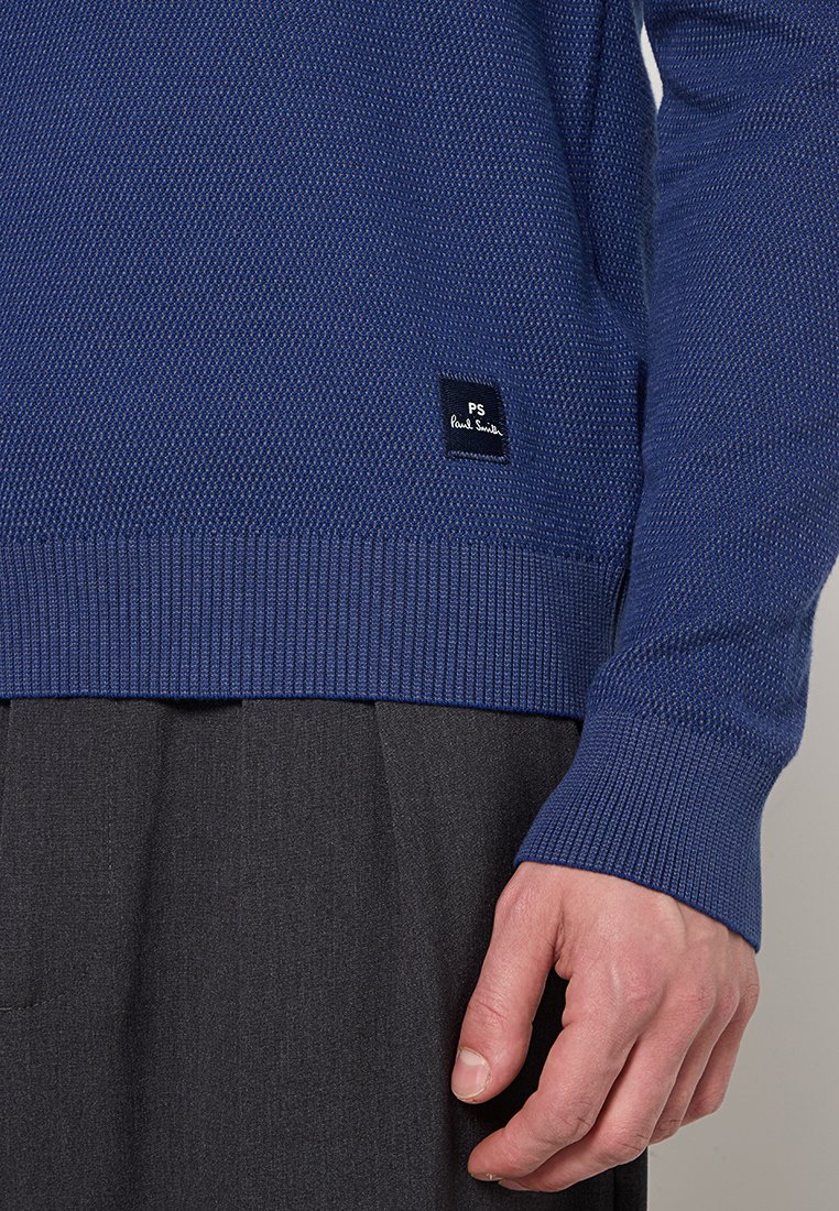 Blå stickad tröja med en strukturerad design, ribbade ärmslut och nederkant, samt en liten vävd etikett med texten "PS Paul Smith."