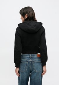 Sudadera negra recortada con capucha, con una textura suave. Combinada con vaqueros de talle alto en azul claro que muestran los bolsillos traseros y una etiqueta de cuero.