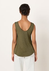 Donna con capelli ricci scuri raccolti in uno chignon, che indossa una canottiera verde oliva e pantaloni chiari, vista da dietro su uno sfondo uniforme.