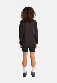 Person mit lockigem Haar steht mit dem Rücken, trägt ein schwarzes langärmliges Oberteil, schwarze Shorts, weiße Socken mit Aufschrift und schwarze Turnschuhe.