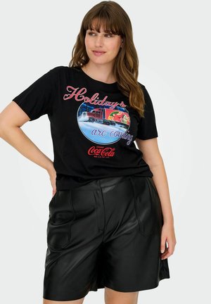 Femme portant un t-shirt noir Coca-Cola « Holidays are coming » avec un motif de camion thème des fêtes et un short en cuir noir.