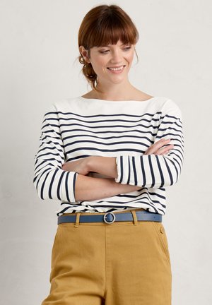 Maglietta a maniche lunghe a righe bianca e blu navy, con scollo a barca. Abbinata a pantaloni color senape e una cintura blu con fibbia circolare.