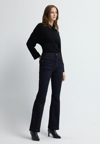 Musta pitkähihaine paita, jossa on kerättynä vyötärö, yhdistettynä tummansinisiin leveneviin farkkuihin, joissa on kontrastivärinen ompelu ja etutaskut.